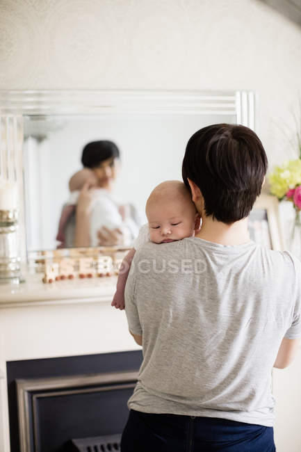 Mãe carregando seu bebê e de pé na frente do espelho em casa — Fotografia de Stock