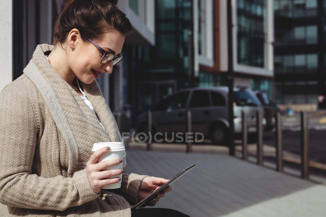 Усміхнена жінка використовує цифровий планшет, тримаючи одноразову чашку на тротуарі — стокове фото