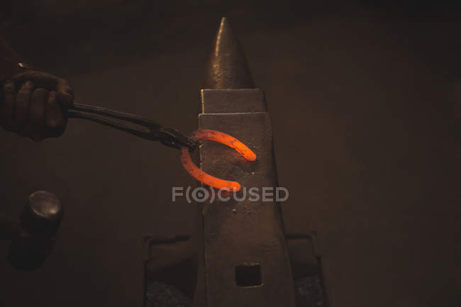 Primo piano del fabbro che tiene il ferro di cavallo di metallo caldo rosso con pinze su incudine in officina — Foto stock