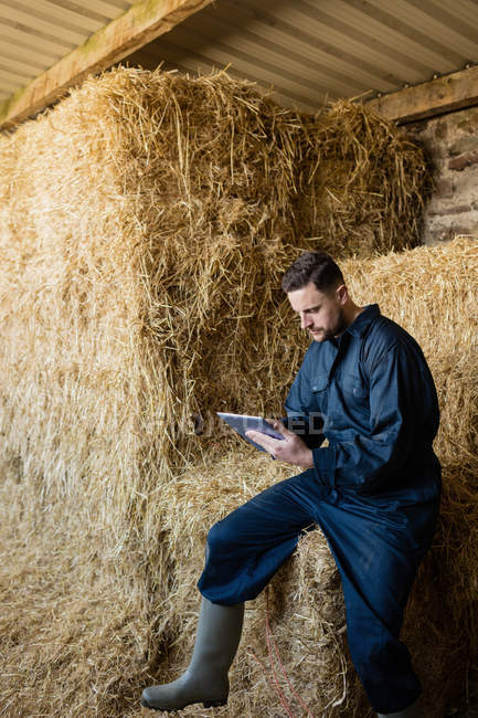 Jeune agriculteur utilisant une tablette numérique assis sur une balle de foin dans une grange — Photo de stock