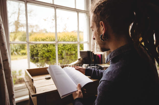 Hipster leyendo novela por ventana en casa - foto de stock
