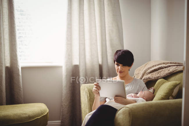 Mère utilisant une tablette numérique pendant que bébé dort dans son bras au salon — Photo de stock