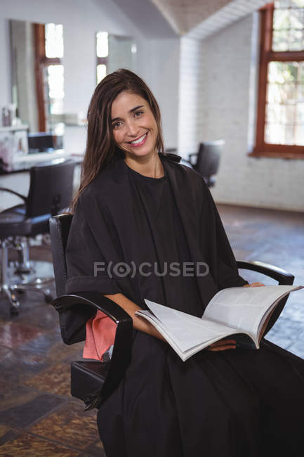 Retrato de una mujer sonriente sentada en una silla y leyendo una revista en un salón de belleza - foto de stock
