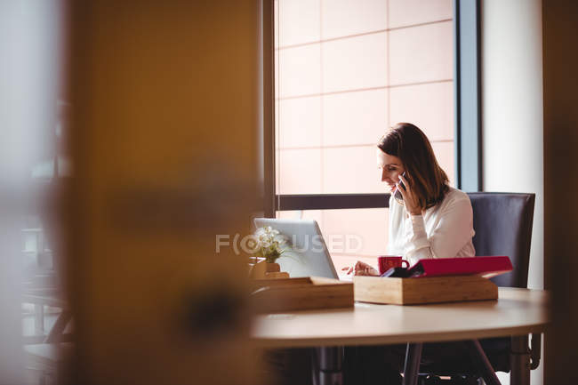 Mujer de negocios usando el ordenador portátil y hablando en el teléfono móvil en la oficina - foto de stock