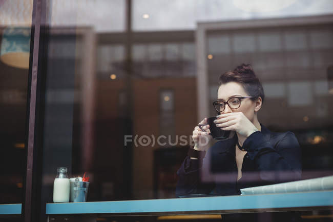 Продумана молода жінка п'є каву в кафе, яку видно через скло — стокове фото