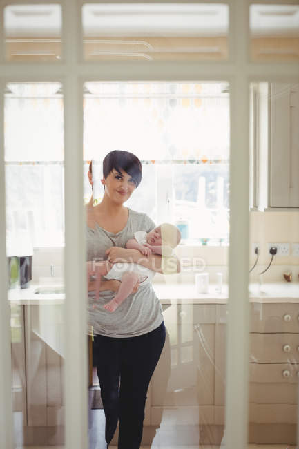 Retrato de la madre sosteniendo a su pequeño bebé en la cocina en casa - foto de stock