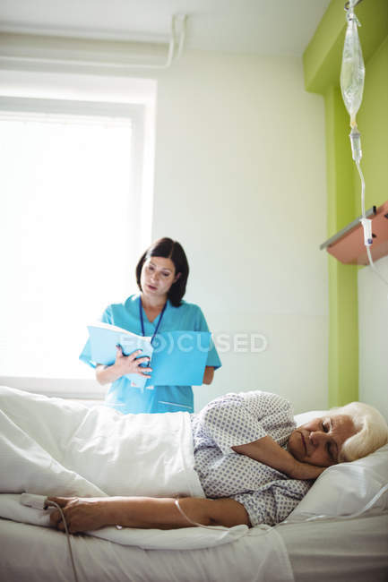 Seniorin schläft auf einem Bett, während Krankenschwester Bericht im Krankenhaus überprüft — Stockfoto
