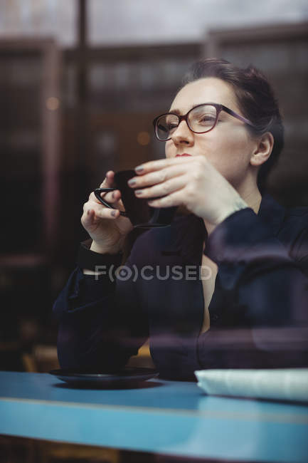 Woman drinking coffee in cafe seen through glass — Stock Photo