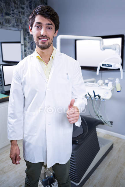 Retrato del dentista sonriente mostrando sus pulgares hacia arriba en la clínica dental - foto de stock