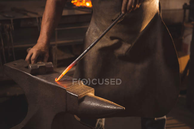 Forgeron mi-section travaillant sur métal chaud à l'aide d'un marteau pour façonner à l'atelier — Photo de stock