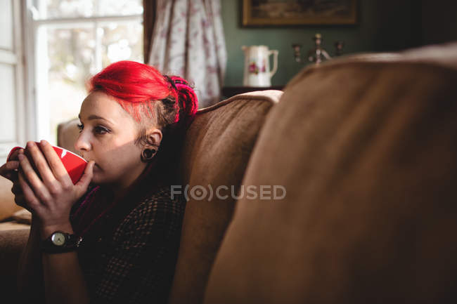 Schöne Frau trinkt Kaffee, während sie zu Hause auf dem Sofa sitzt — Stockfoto