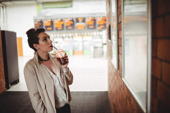 Young woman looking towards board while sipping drink at railroad station — Stock Photo