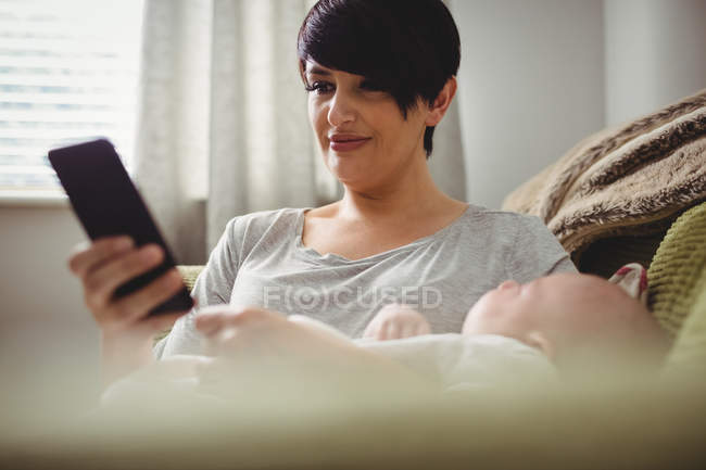 Madre usando teléfono inteligente mientras el bebé duerme en su brazo en la sala de estar - foto de stock