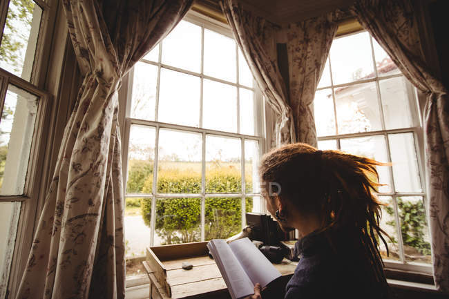 Vista laterale del romanzo di lettura hipster a tavola per finestra — Foto stock