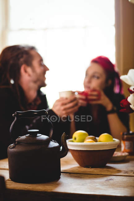 Bule e maçãs na tigela na mesa com casal no fundo — Fotografia de Stock