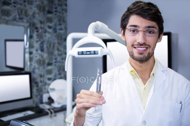 Retrato de dentista sorridente em pé com uma ferramenta dentária na clínica odontológica — Fotografia de Stock
