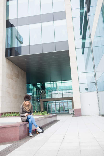 Junge Frau benutzt digitales Tablet, während sie vor Bürogebäude sitzt — Stockfoto