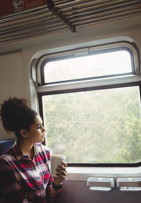 Mujer pensativa mirando a través de la ventana mientras toma café en tren - foto de stock