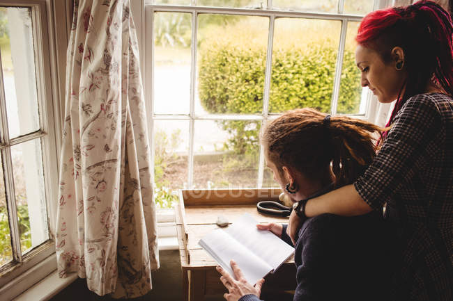 Vista lateral do jovem casal leitura livro por janela em casa — Fotografia de Stock