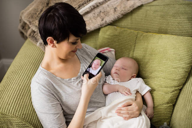Mother taking picture of her baby with smartphone in living room at home — Stock Photo