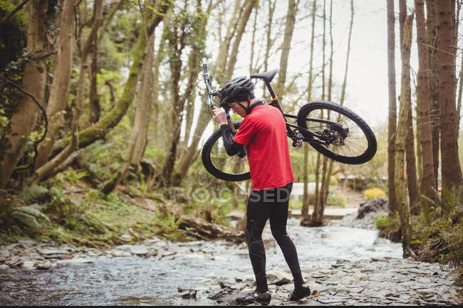 Biciclista de montanha carregando bicicleta enquanto atravessa o riacho na floresta — Fotografia de Stock