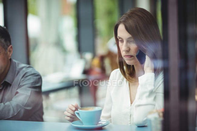 Женщина держит чашку кофе и разговаривает по мобильному телефону в кафетерии — стоковое фото
