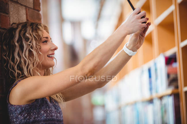 Красивая женщина делает селфи с мобильным телефоном в библиотеке — стоковое фото