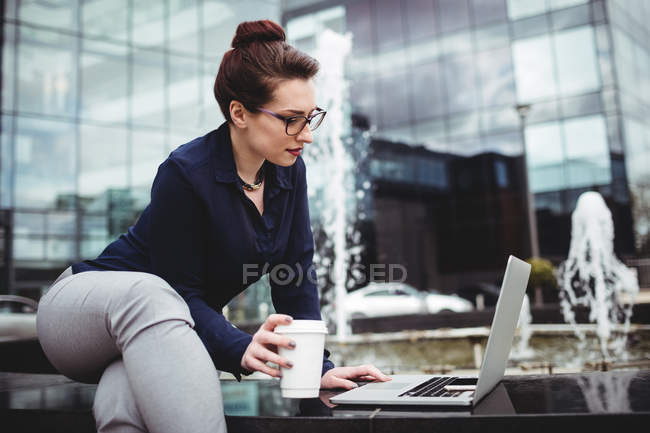 Joven mujer de negocios utilizando el ordenador portátil contra el edificio de oficinas moderno - foto de stock