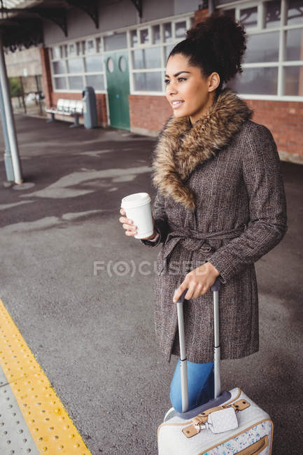 Улыбающаяся женщина с одноразовой чашкой кофе, стоя на платформе железнодорожного вокзала — стоковое фото