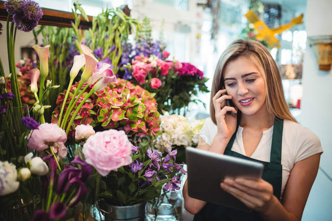 Female florist using digital tablet while talking on mobile phone in the flower shop — Stock Photo