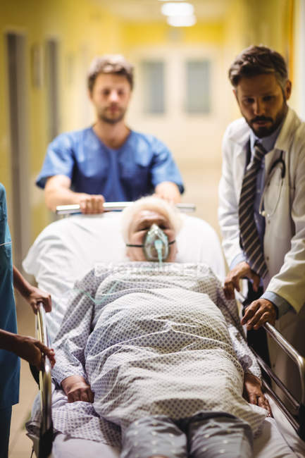 Médico y niño de sala empujando paciente mayor en camilla en el pasillo del hospital - foto de stock