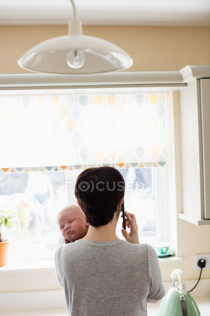 Вид сзади: мать держит своего маленького ребенка, разговаривая по смартфону на кухне дома — стоковое фото