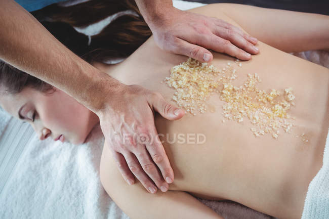 Fisioterapeuta aplicando exfoliante de sal en la espalda de una paciente femenina en la clínica - foto de stock