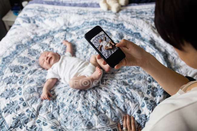 Mother taking picture of her baby by smartphone in bedroom at home — Stock Photo