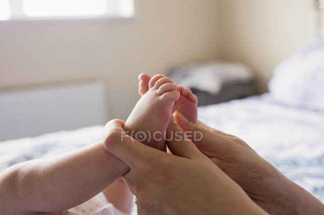 Imagen recortada de la madre sosteniendo los pies del bebé en la cama en casa - foto de stock