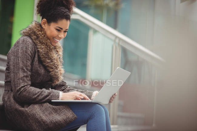 Усміхнена красива жінка використовує ноутбук, сидячи на сходах — стокове фото