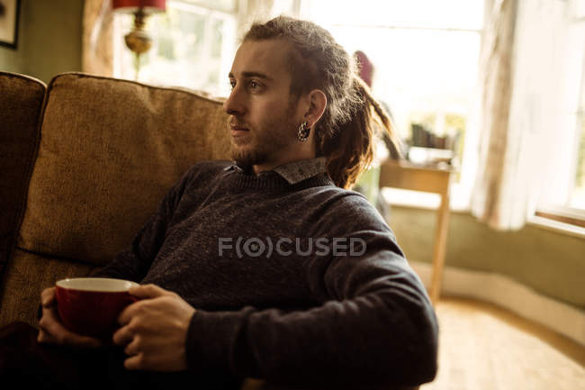 Vista laterale dell'hipster che regge la tazza di caffè mentre si rilassa sul divano di casa — Foto stock