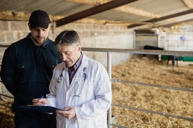 Vet discussing with farm worker over clipboard by fence in barn — Stock Photo