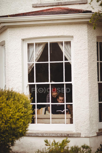 Пара читає книгу в будинку, яку видно через вікно — стокове фото