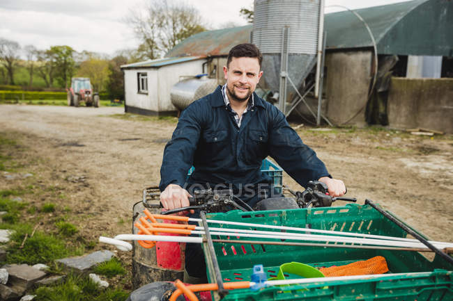 Retrato de jovem agricultor montando quadriciclo em campo contra celeiro — Fotografia de Stock