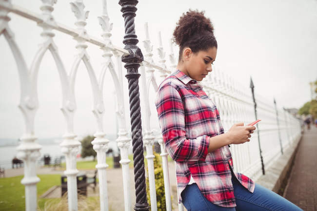 Вид сбоку на молодую женщину, которая использует телефон, опираясь на перила — стоковое фото