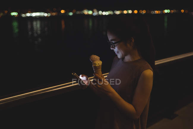 Mujer joven usando el teléfono móvil mientras toma helado por la noche - foto de stock