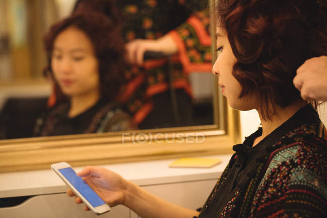 Donna che utilizza il telefono cellulare mentre ottenere i capelli raddrizzati a parrucchiere — Foto stock