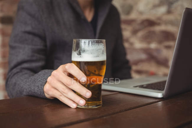Homem segurando copo de cerveja e usando laptop no bar — Fotografia de Stock