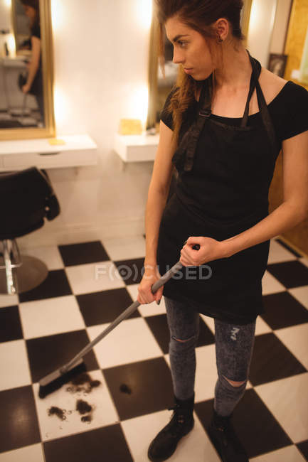 Femme salon de coiffure nettoyage des déchets de cheveux sur le sol avec balai au salon — Photo de stock