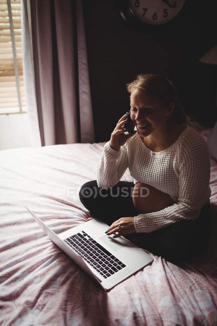 Беременная женщина разговаривает на смартфоне во время использования ноутбука в спальне дома — стоковое фото