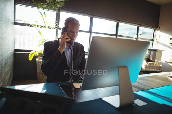 Businessman talking on mobile phone while using desktop pc in office — Stock Photo