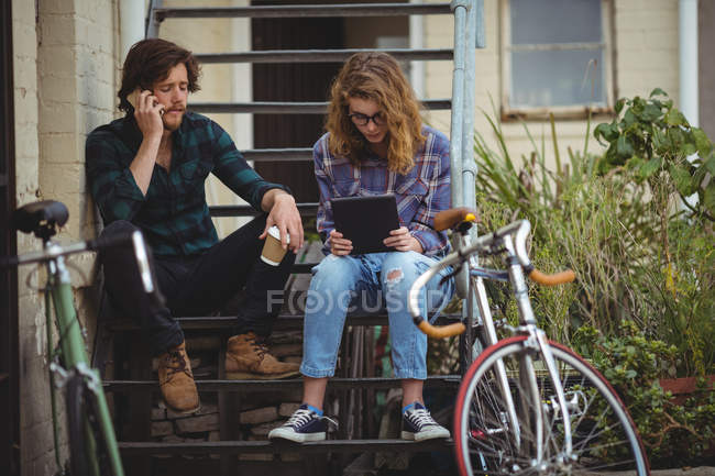Paar sitzt auf Treppe, während es mit dem Handy telefoniert und ein digitales Tablet nutzt — Stockfoto