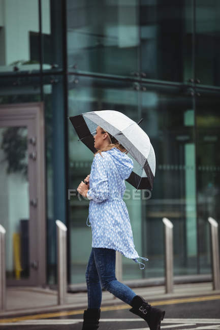 Vista lateral da bela mulher segurando guarda-chuva e andando na rua durante a estação chuvosa — Fotografia de Stock