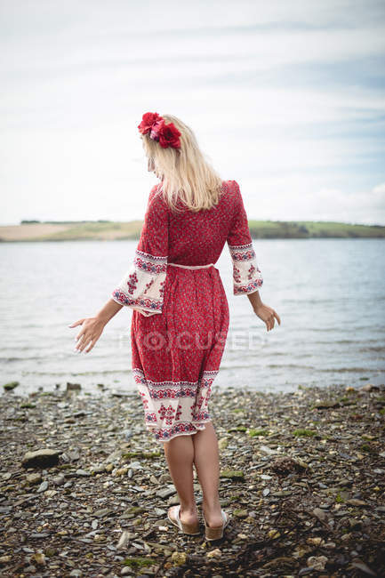 Vista trasera de la mujer rubia despreocupada en vestido rojo y tiara de flores de pie cerca del río - foto de stock